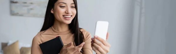 Mujer asiática feliz con portátil apuntando con el dedo durante el chat de vídeo en el teléfono inteligente, bandera - foto de stock