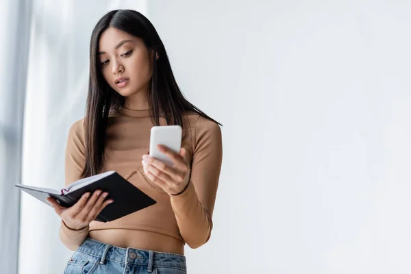 Asian woman holding notebook while looking at cellphone — Stock Photo