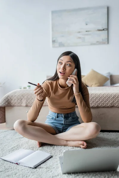 Excited asian woman gesturing with pen while talking on smartphone on floor near blurred laptop — Stock Photo