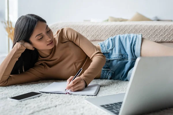 Young asian woman writing in notebook while lying on floor near blurred laptop — Stock Photo