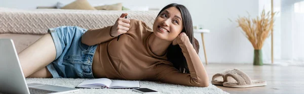 Cheerful asian woman holding pen while lying on floor near gadgets and notebook, banner — Stock Photo