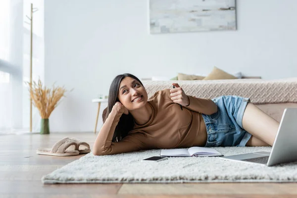Sonriendo mujer asiática acostada en la alfombra en el dormitorio cerca de gadgets y portátil - foto de stock