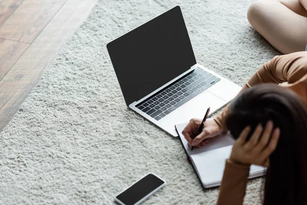 Vista aérea de la mujer borrosa escribiendo en el portátil cerca de la computadora portátil y el teléfono inteligente en la alfombra del piso - foto de stock