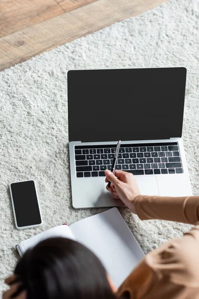 Overhead view of blurred woman pointing with pen at laptop near smartphone with blank screen on floor — Stock Photo