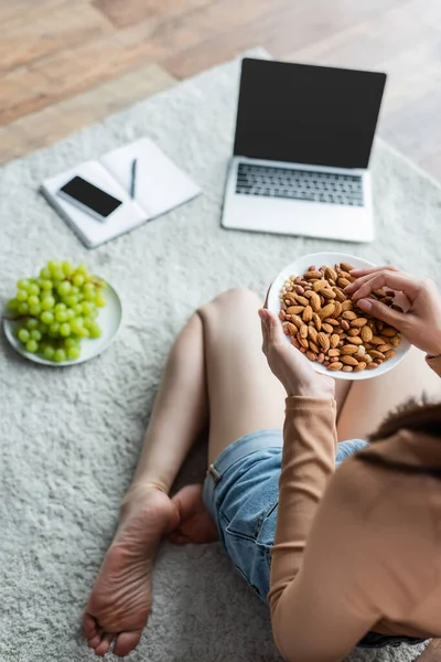 Vue recadrée du pigiste avec bol d'amandes assis sur le sol près des gadgets et du raisin frais — Photo de stock