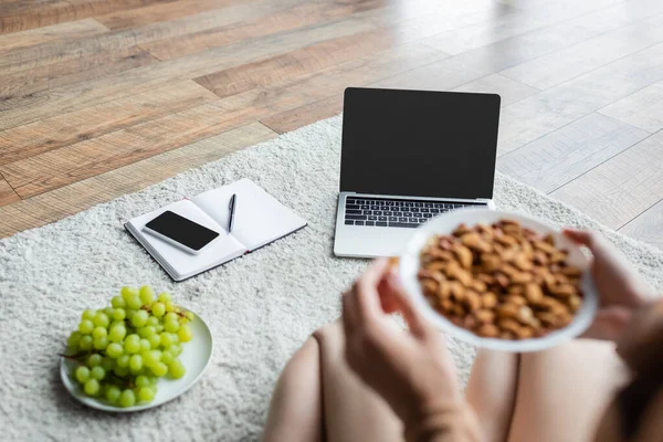 Teilansicht eines verschwommenen Freelancer, der Schüssel mit Mandeln in der Nähe von Gadgets und frischer Traube auf dem Boden hält — Stockfoto