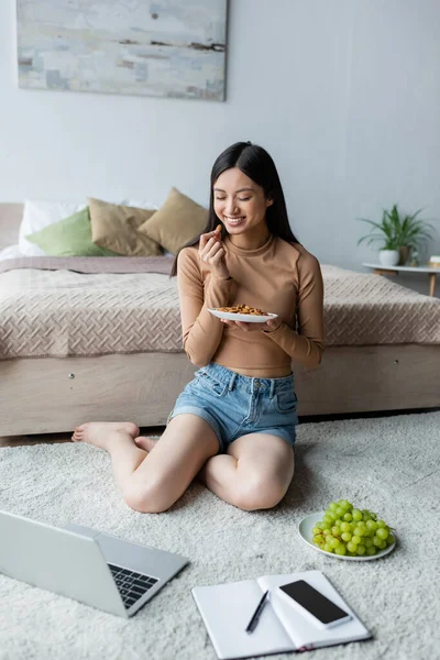 Feliz asiático freelancer comer amêndoas perto laptop e lanches no chão no quarto — Fotografia de Stock