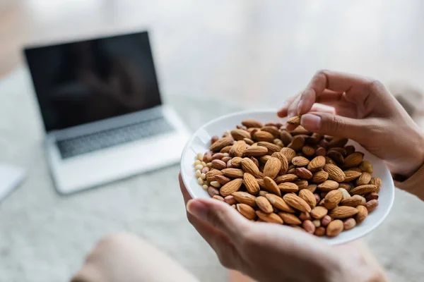 Partial view of woman with bowl of almonds near blurred laptop with blank screen — Stock Photo