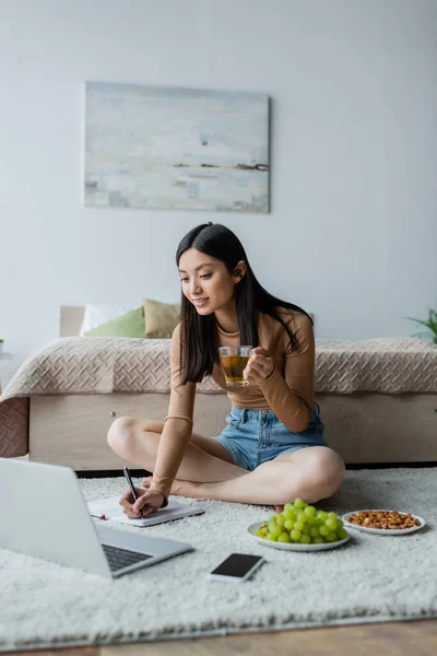 Mulher asiática com xícara de chá escrevendo em notebook perto de gadgets, amêndoas e uva no chão — Fotografia de Stock