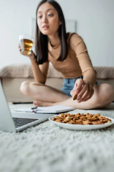 Verschwommene asiatische Frau, die Tee trinkt und Mandeln isst, während sie auf den Laptop am Boden schaut — Stockfoto