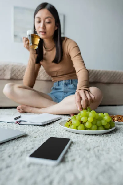 Desfocado asiático mulher tomando uva enquanto bebe chá no chão perto notebook e smartphone — Fotografia de Stock