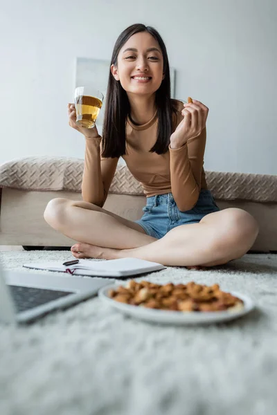 Feliz asiático mulher comer amêndoas e beber chá no chão perto turvo laptop — Fotografia de Stock