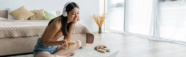 Mujer asiática feliz en los auriculares sentados en el suelo con la taza de té cerca de la computadora portátil, bandera - foto de stock