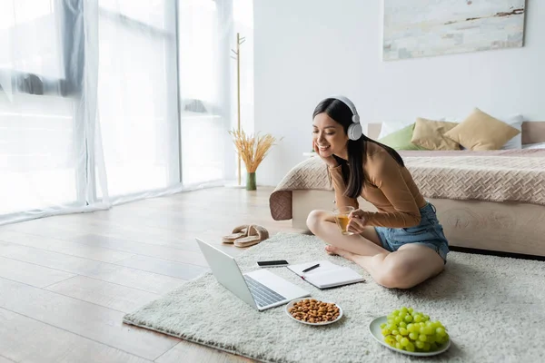 Glückliche asiatische Frau mit Kopfhörern sitzt auf dem Boden mit einer Tasse Tee in der Nähe von Gadgets und Snacks — Stockfoto