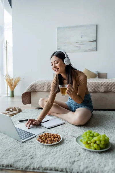 Smiling asian woman with cup of tea using laptop on floor in bedroom — Stock Photo