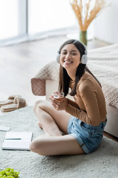 Fröhliche asiatische Frau mit Kopfhörern, die eine Tasse Tee hält, während sie mit überkreuzten Beinen neben dem Notizbuch auf dem Boden sitzt — Stockfoto
