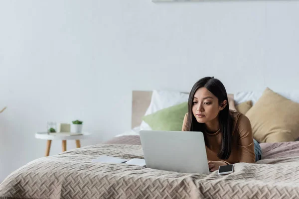 Brunette asian woman using laptop while lying on bed at home — Stock Photo