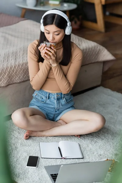 Asian woman in headphones drinking tea near laptop and notebook on floor — Stock Photo