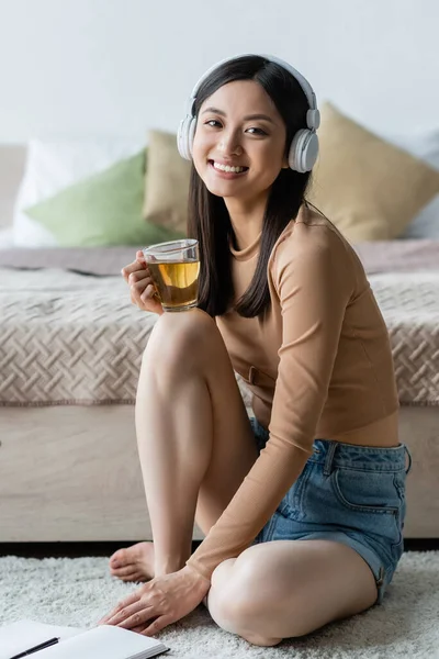 Азиатка в наушниках с чашкой чая и улыбкой на камеру в спальне — стоковое фото