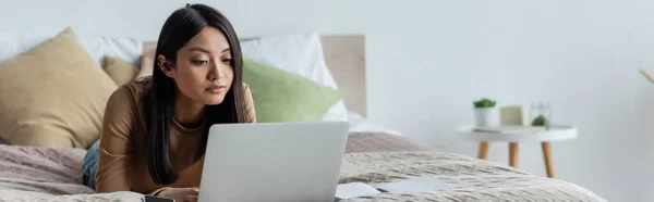 Junge Asiatin arbeitet am Laptop, während sie zu Hause auf dem Bett liegt, Banner — Stockfoto
