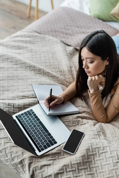 Hochwinkelaufnahme einer Asiatin, die in einem Notizbuch in der Nähe von Laptop und Smartphone auf dem Bett schreibt — Stockfoto