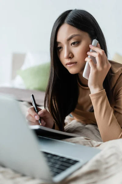 Asiatin schaut auf verschwommenen Laptop, während sie auf dem Bett mit dem Handy spricht — Stockfoto