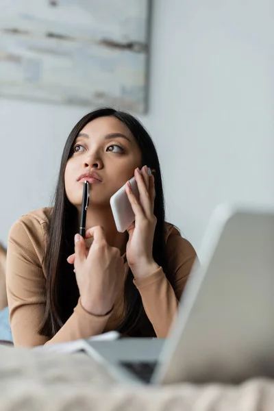 Premuroso asiatico freelancer parlando su cellulare vicino offuscata laptop in camera da letto — Foto stock