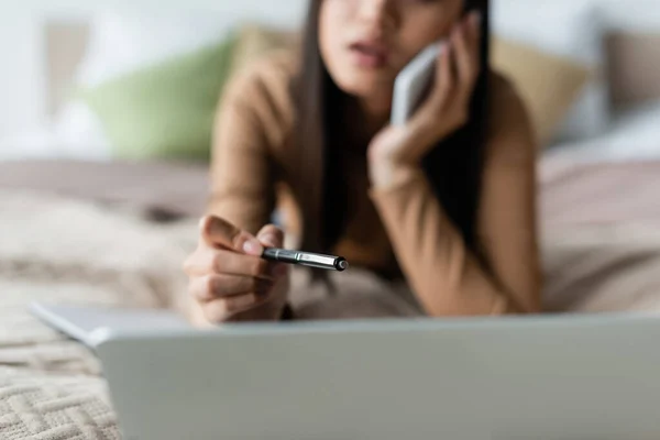 Mujer borrosa que apunta con la pluma en el ordenador portátil mientras habla en el teléfono celular en el dormitorio - foto de stock