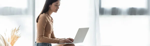 Pretty asian freelancer typing on laptop near window at home, banner — Stock Photo