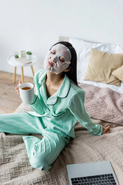 Asiático mujer en hoja máscara sentado en cama con taza de té y cerrado ojos cerca de laptop - foto de stock