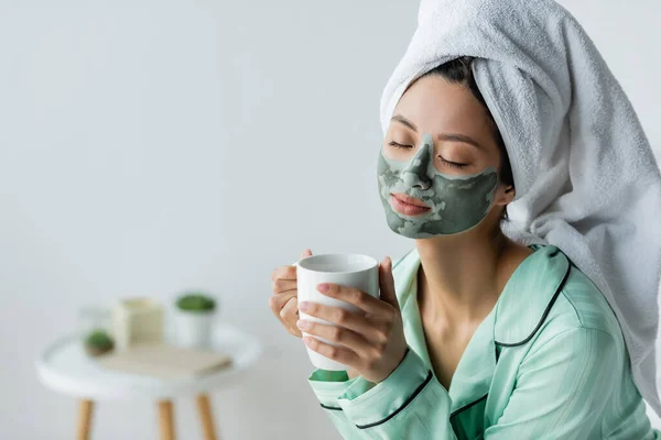 Heureux asiatique femme en argile masque, pyjama et serviette sur la tête tenant tasse de thé avec les yeux fermés — Photo de stock