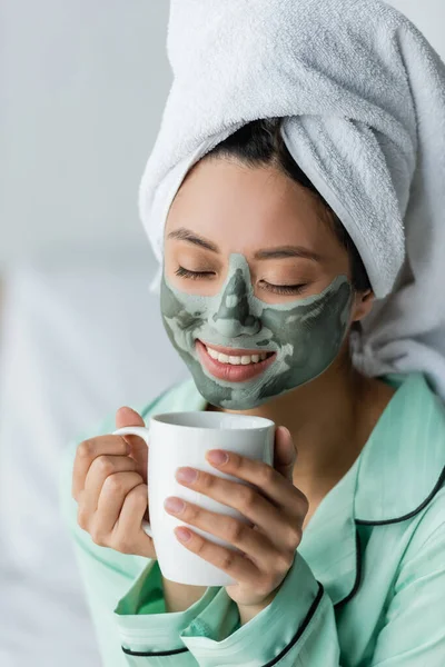 Joyful asian woman with clay mask on face and towel on head drinking tea with closed eyes — Stock Photo