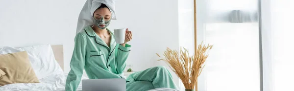 Donna asiatica in maschera di argilla e pigiama seduta sul letto con tazza di tè vicino al computer portatile, banner — Foto stock