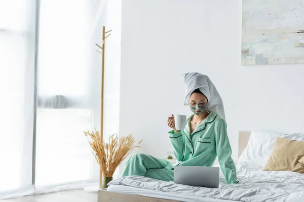 Mulher asiática em máscara de barro e pijama sentado na cama com xícara de chá e olhando para o laptop — Fotografia de Stock