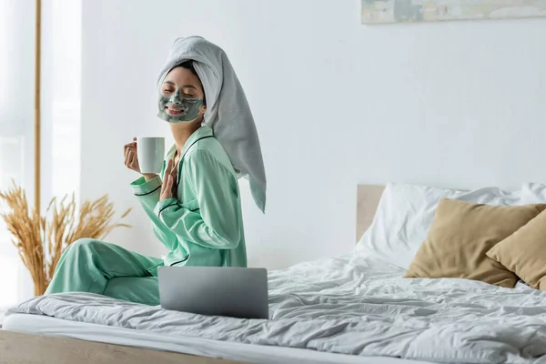 Mujer asiática complacida con los ojos cerrados sosteniendo la taza de té mientras está sentado cerca del ordenador portátil en el dormitorio - foto de stock