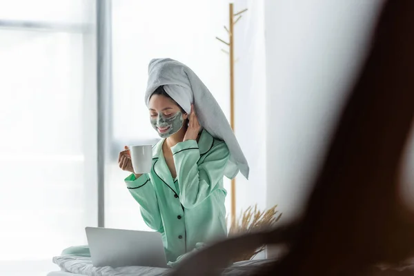 Smiling asian woman in pajamas and clay mask sitting with cup of tea near laptop in bedroom — Stock Photo