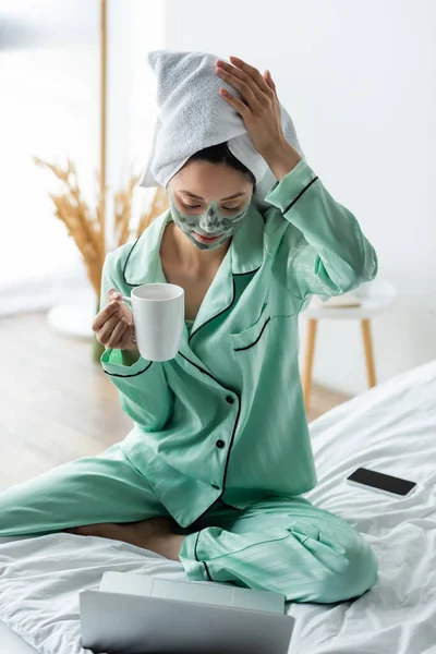 Asiatische Frau in Lehmmaske und Handtuch auf dem Kopf sitzt auf dem Bett mit einer Tasse Tee in der Nähe von Laptop — Stockfoto