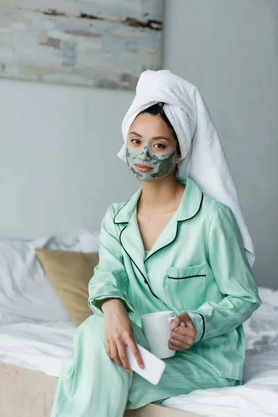 Donna asiatica in pigiama e maschera di argilla seduta sul letto con smartphone e tazza di tè — Foto stock