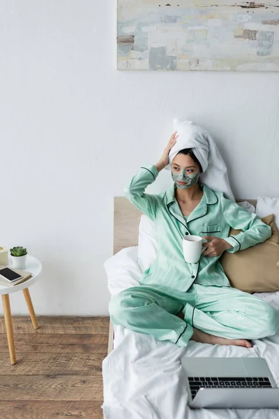 Азиатка в глиняной маске и пижаме трогает полотенце на голове, сидя рядом с ноутбуком на кровати — стоковое фото