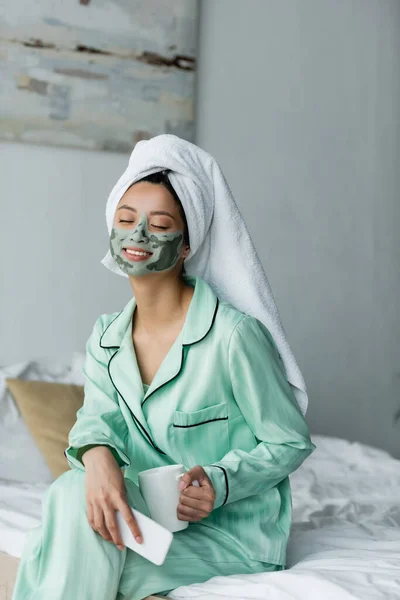 Довольная азиатка с глиняной маской на лице сидит на кровати с мобильным телефоном и чаем — стоковое фото