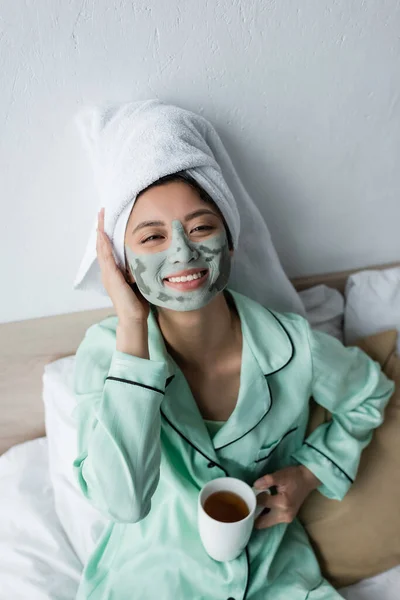 Mujer asiática feliz con toalla en la cabeza y máscara de arcilla en la cara sosteniendo taza de té mientras sonríe a la cámara - foto de stock