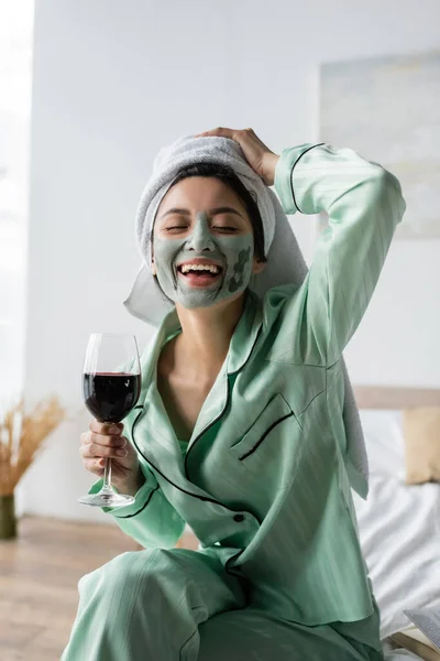 Eccitato donna asiatica in maschera di argilla e asciugamano sulla testa in possesso di un bicchiere di vino rosso e ridere con gli occhi chiusi — Foto stock