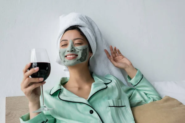 Happy asian woman in pajamas, towel on head and clay mask holding red wine in bed — Stock Photo