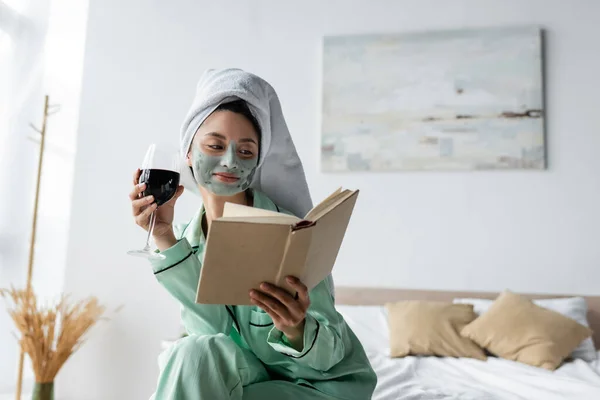 Smiling asian woman in pajamas and clay mask holding red wine while reading book in bedroom — Stock Photo