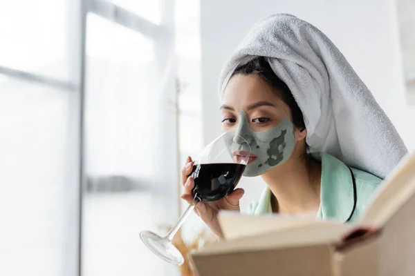 Asian woman with clay mask and towel on head drinking red wine while reading book at home — Stock Photo