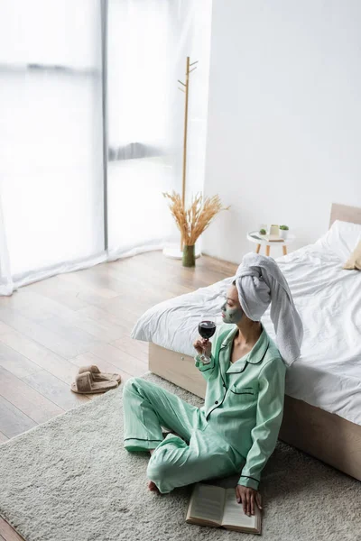 Hochwinkel-Ansicht der asiatischen Frau im Pyjama und Lehmmaske sitzt auf dem Boden im Schlafzimmer mit einem Glas Rotwein — Stockfoto