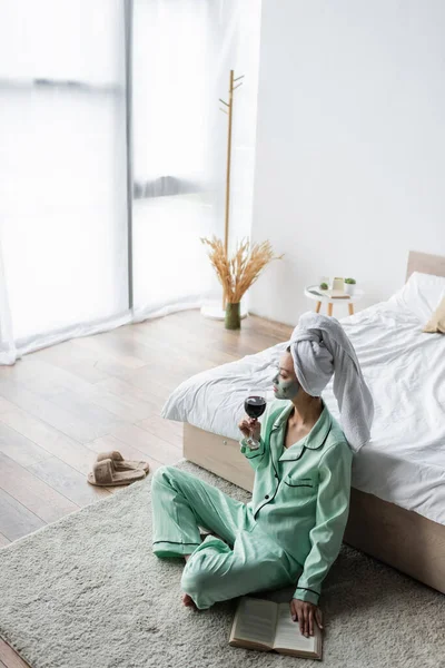 Vue grand angle de la femme asiatique en pyjama et masque d'argile assis sur le sol dans la chambre avec verre de vin rouge — Photo de stock