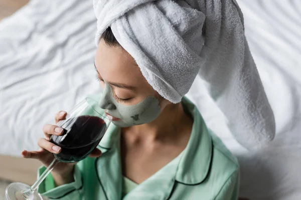 Hochwinkelaufnahme einer asiatischen Frau in Lehmmaske und Handtuch auf dem Kopf, die im Schlafzimmer Rotwein trinkt — Stockfoto