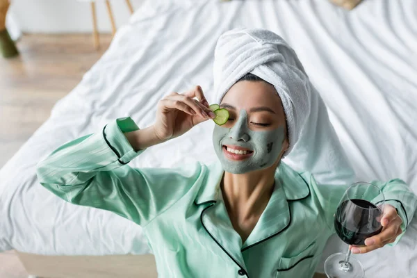 Freudige asiatische Frau in Tonmaske, die Gurkenscheiben auf das Auge aufträgt, während sie ein Glas Rotwein in der Hand hält — Stockfoto