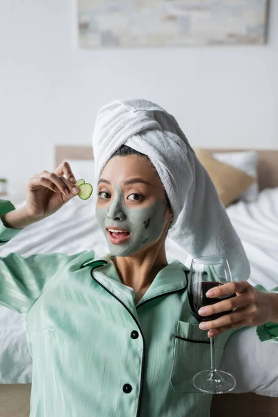 Joven asiático mujer en arcilla máscara y pijama celebración pepino rebanadas y vaso de vino tinto mientras mira cámara - foto de stock